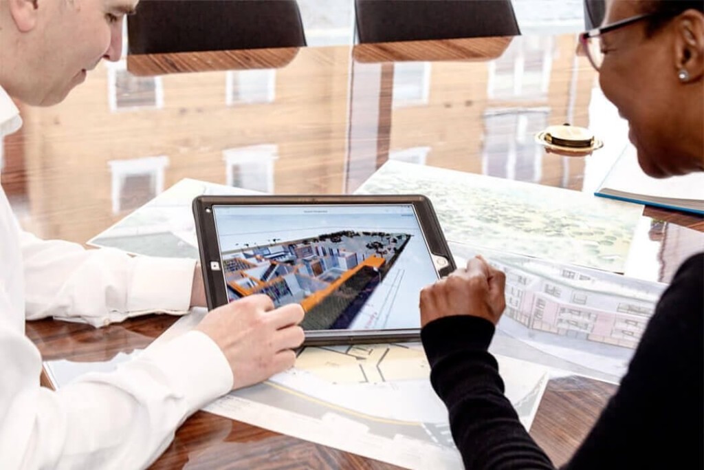 Two architects collaborate on a modern housing project using a digital tablet. The screen displays a detailed 3D model of an urban development, surrounded by blueprints and architectural plans on a polished wooden table. The scene highlights the importance of advanced planning, architectural design, and professional consultation in achieving successful property extensions.
