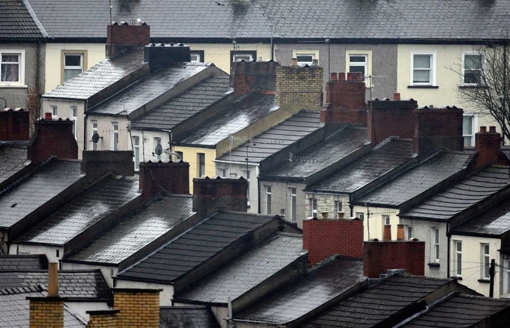 An aerial view of a dense row of terraced houses in an urban area. The rooftops are wet from rain, and chimneys and satellite dishes are visible on many of the houses. The image highlights the architectural uniformity and compact living conditions common in older urban neighbourhoods. This photograph is an example of housing stock that could benefit from upward extensions to increase living space without expanding the building footprint.
