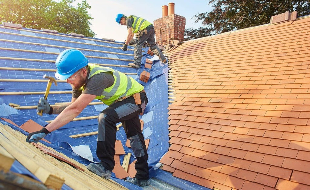 Construction workers are actively engaged in a roofing project, meticulously aligning and securing new tiles atop a house. Clad in safety gear, including blue helmets and reflective vests, they work on both the newly constructed and existing roof sections. The bright sunlight and clear sky suggest ideal working conditions.