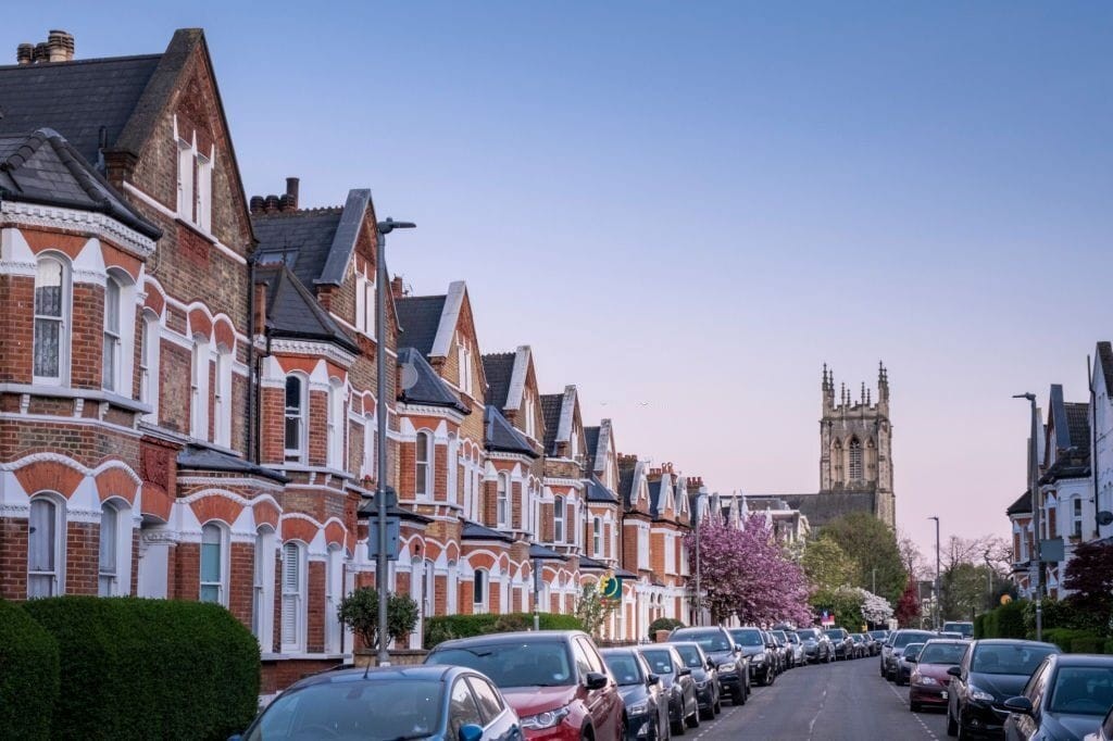 A picturesque row of Victorian terraced houses on a quiet London street at dusk, showcasing the characteristic red brick facades, bay windows, and steep slate roofs. A historic church tower is visible in the background, adding to the charming architectural scene.