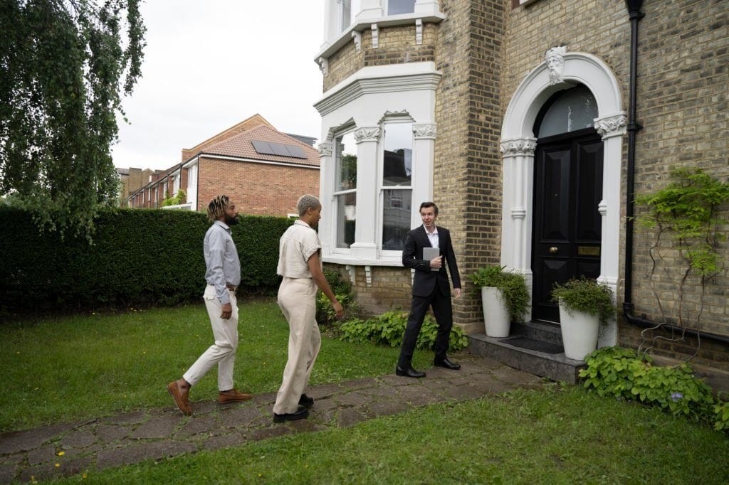 A real estate agent leading two potential buyers towards the entrance of a Victorian house with a well-maintained garden. The house features traditional brickwork, ornate window trims, and a black front door with arched detailing, capturing the classic architectural elements of Victorian homes. The lush green lawn and hedges enhance the curb appeal of the property.
