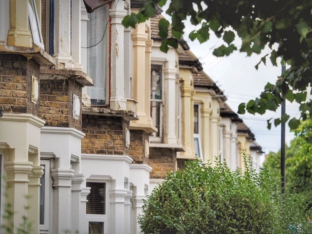 A row of Victorian terraced houses in a leafy neighborhood, highlighting the distinctive architectural features such as ornate cornices, large bay windows, and detailed brickwork. The houses exhibit a mix of restored and weathered facades, showcasing the charm and character of Victorian-era design.