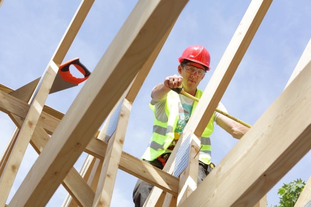 Construction worker in a red hard hat and high-visibility vest measures timber framing with a tape measure on a sunny day. He is part of a team working on extending and refurbishing a Victorian house. The image captures the early stages of the build, highlighting the structural framework and the use of modern construction techniques to renovate historical architecture. The blue sky provides a clear backdrop, emphasising the progress and craftsmanship involved in the project.