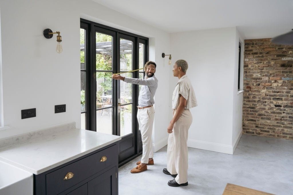 A couple viewing the interior of a refurbished Victorian house, featuring modern updates with traditional charm. They stand by large black-framed glass doors leading to a garden, while discussing potential renovations. The room showcases exposed brickwork, a sleek black kitchen island with brass handles, and a white marble countertop, blending contemporary design with classic Victorian elements. The space is well-lit with natural light, enhancing the airy and open feel.