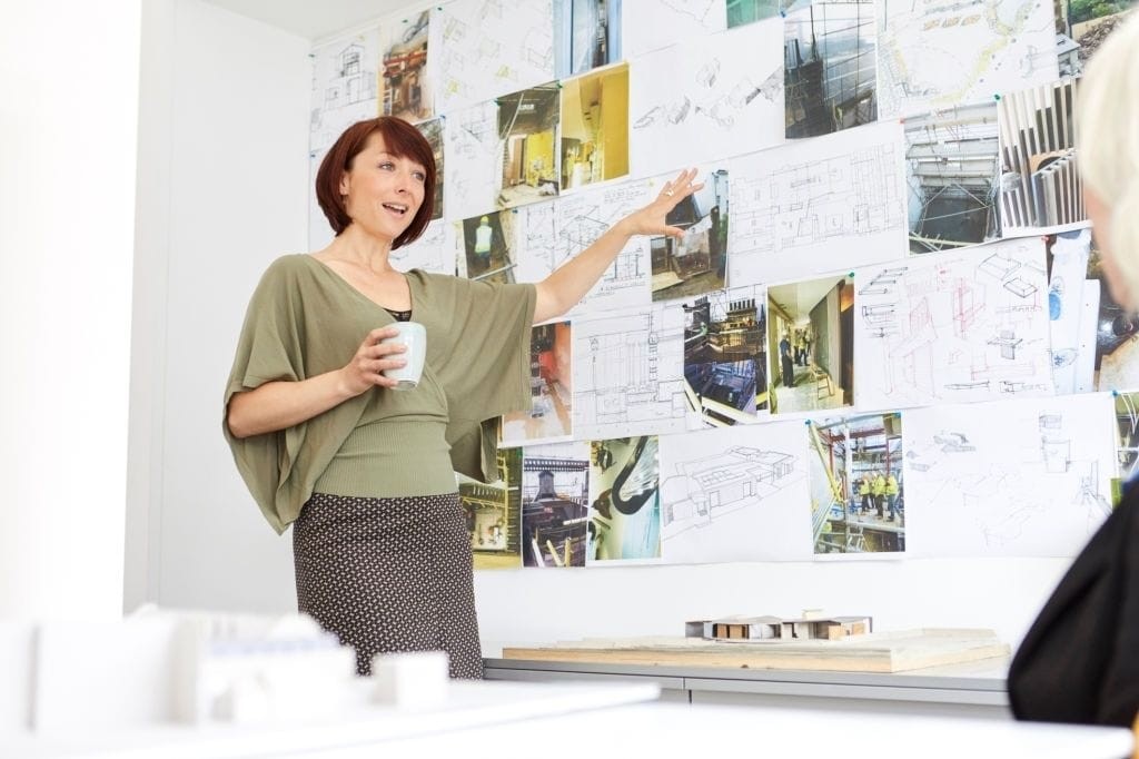 Female architect presenting urban development plans with a coffee mug in hand, gesturing towards project images and blueprints pinned on the wall, with architectural models on the table, in a bright office setting.