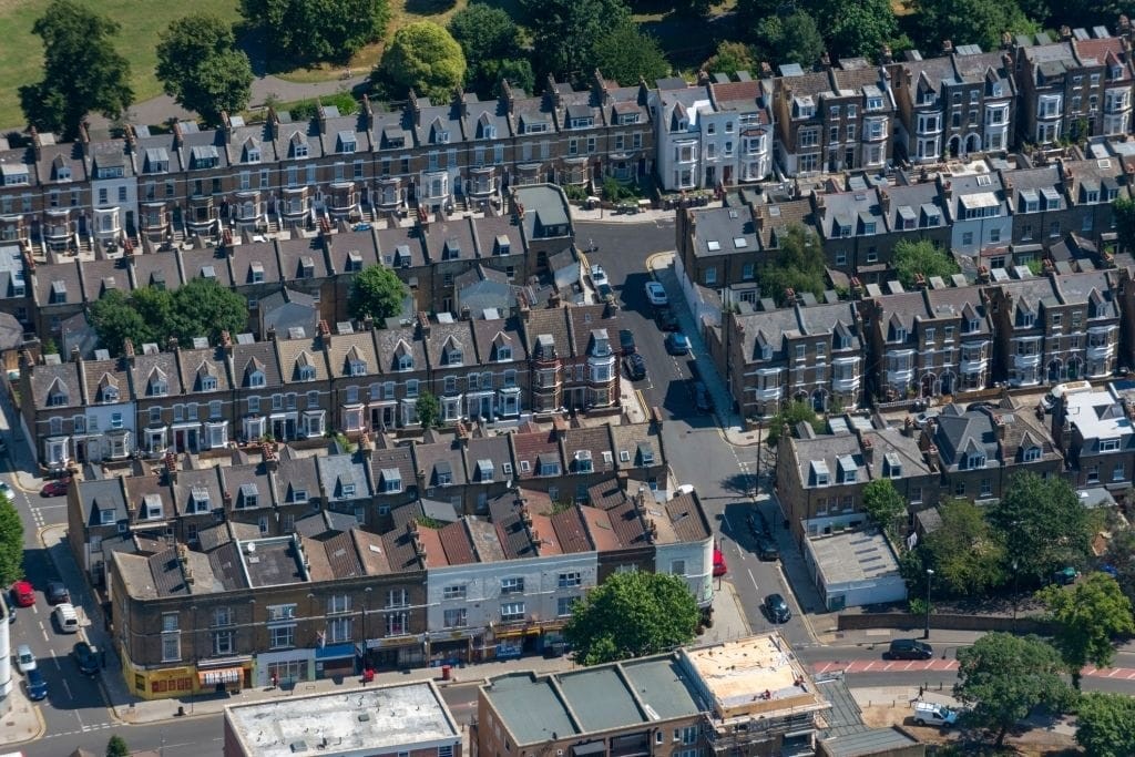Aerial view of a dense residential area in London, showcasing rows of terraced houses with uniform design and gabled roofs, interspersed with trees and a few commercial properties, on a sunny day.