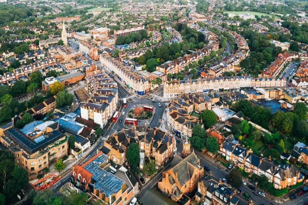 Aerial view of a bustling neighborhood at dawn with historic terraced houses and modern buildings intersected by curving streets and roundabouts, showcasing the vibrant residential and commercial architecture in a suburban area of London.