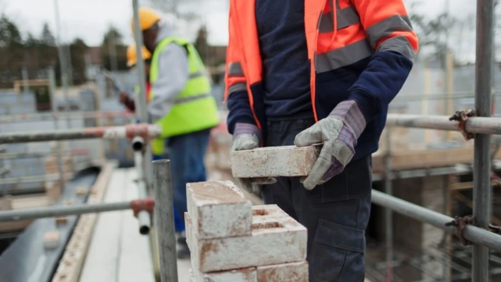 Construction worker in a hi-vis jacket placing a brick on a stack on scaffolding at a London building site, with another worker blurred in the background.