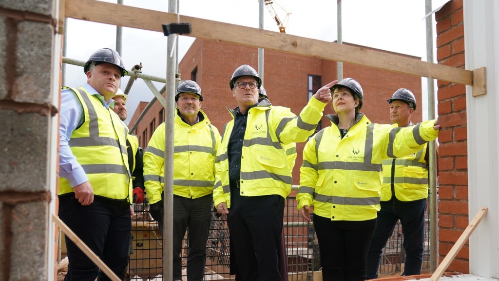 Group of people in high-visibility jackets and hard hats on a construction site, discussing plans for a film studio development in the Green Belt