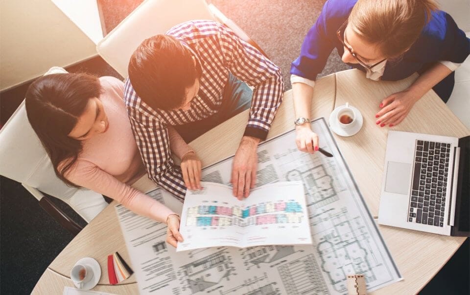 Top-down view of three professionals reviewing architectural plans on a table with a laptop and coffee, indicating a collaborative planning session in an office environment.