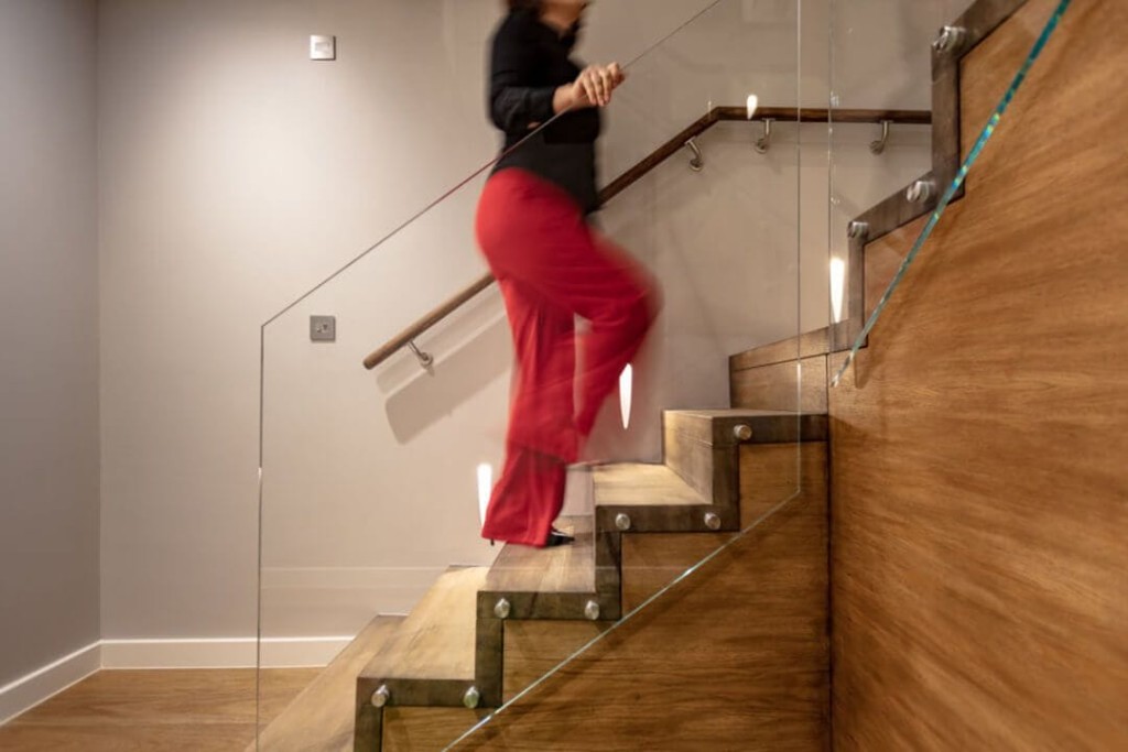Motion blur image of a person in red trousers ascending a wooden staircase with glass balustrade in a modern home interior.
