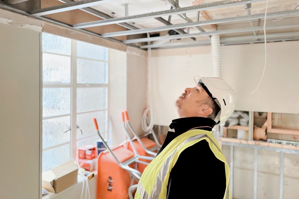 Construction site inspection with a focused engineer wearing a white helmet and high-visibility jacket, looking up at ceiling structures in a room with natural light and construction equipment in the background.