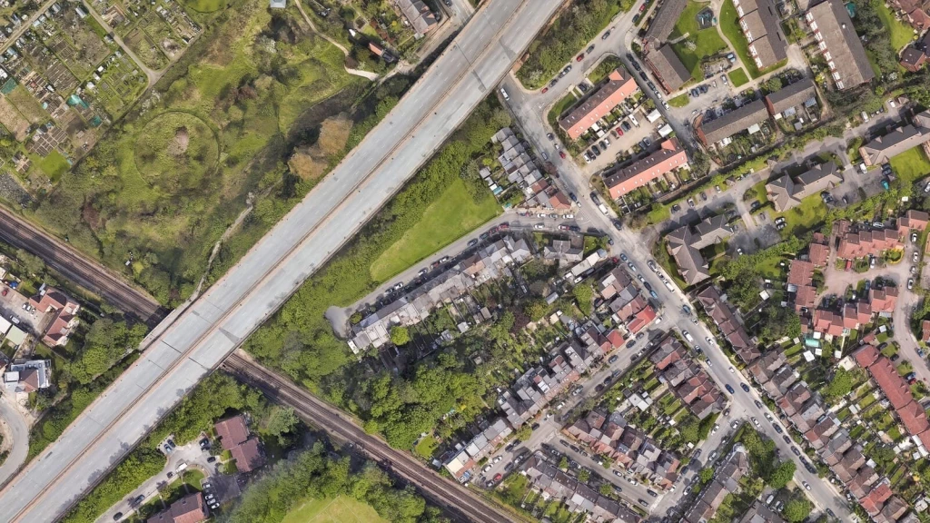 Aerial photograph of a suburban neighbourhood: rows of terraced and semi‑detached houses cluster around narrow streets, while a broad dual carriageway slices diagonally through the scene and pockets of undeveloped greenfield land and allotments lie unused, an illustration of a council failing to meet five‑year housing land supply.