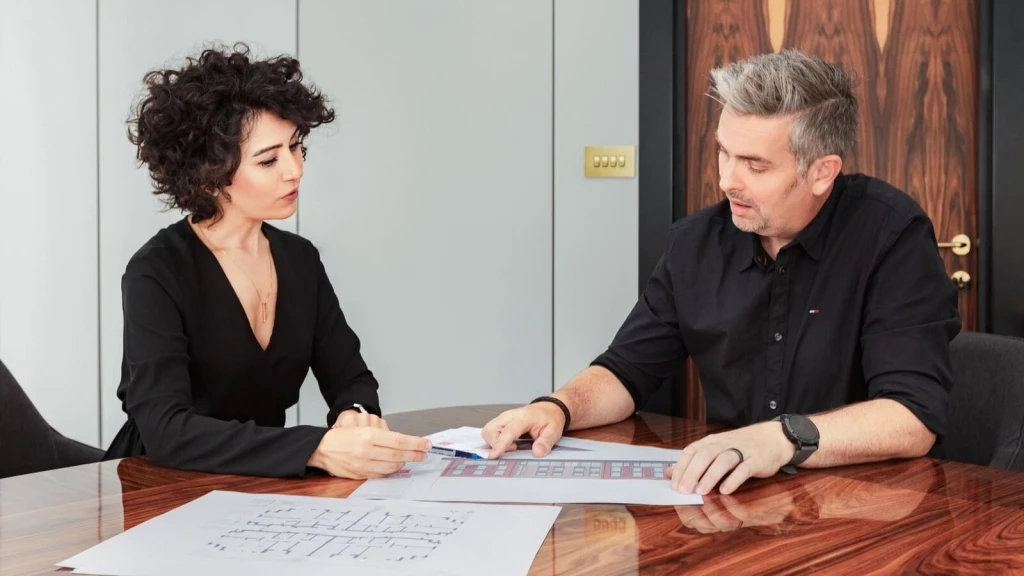 Directors at Urbanist Architecture in a professional meeting discussing building plans. The chartered town planner, with curly hair and dressed in a black blouse, and the chartered architect, with short grey hair and wearing a black shirt, are seated at a polished wooden table with architectural blueprints spread out in front of them. They are deeply engaged in reviewing the designs, symbolising collaboration and expertise in architectural planning.