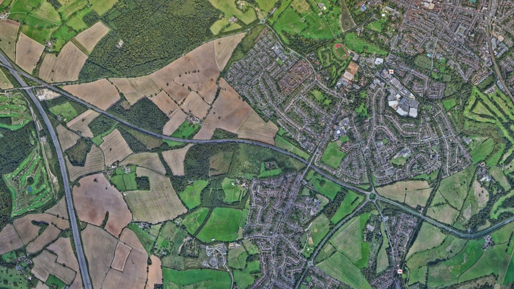 Aerial view of a landscape showing a mix of urban and rural areas, illustrating the boundaries between developed neighbourhoods and large green spaces typical of Green Belt land. Fields, forests, and residential areas are visible, separated by roads and natural features. This image highlights the contrast between protected Green Belt land and adjacent urban sprawl, relevant to discussions on Green Belt boundary reviews, urban expansion, and land release for housing developments.