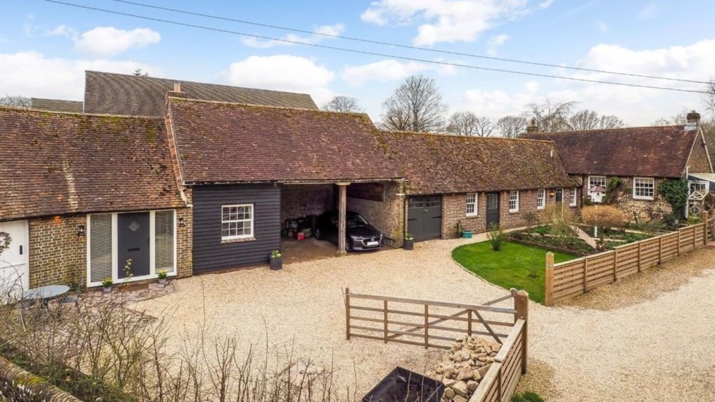 Traditional country cottage with a charming brick and dark timber facade, featuring a gravel driveway, a carport with a parked car, and a well-kept garden, set against a backdrop of mature trees.