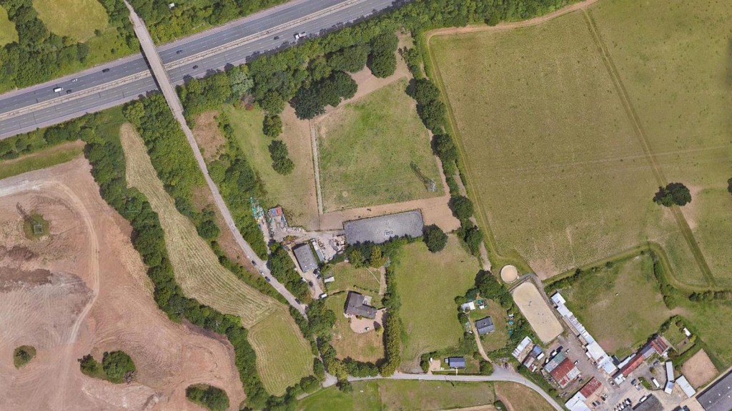 Overhead view of a mixed-use landscape adjacent to a highway, featuring agricultural fields, construction areas, a small cluster of buildings, and patches of dense greenery, illustrating land use variation.