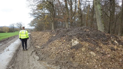 Man in high-visibility jacket walking along a muddy track beside a pile of excavated soil and woodland on Green Belt land, showing how parts of the Green Belt are industrial or disturbed rather than natural countryside.