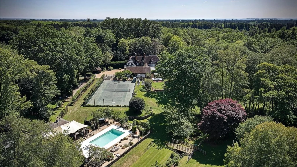Aerial view of a luxury country estate in the Green Belt with Tudor-style manor house, tennis court, swimming pool, and landscaped gardens surrounded by woodland.