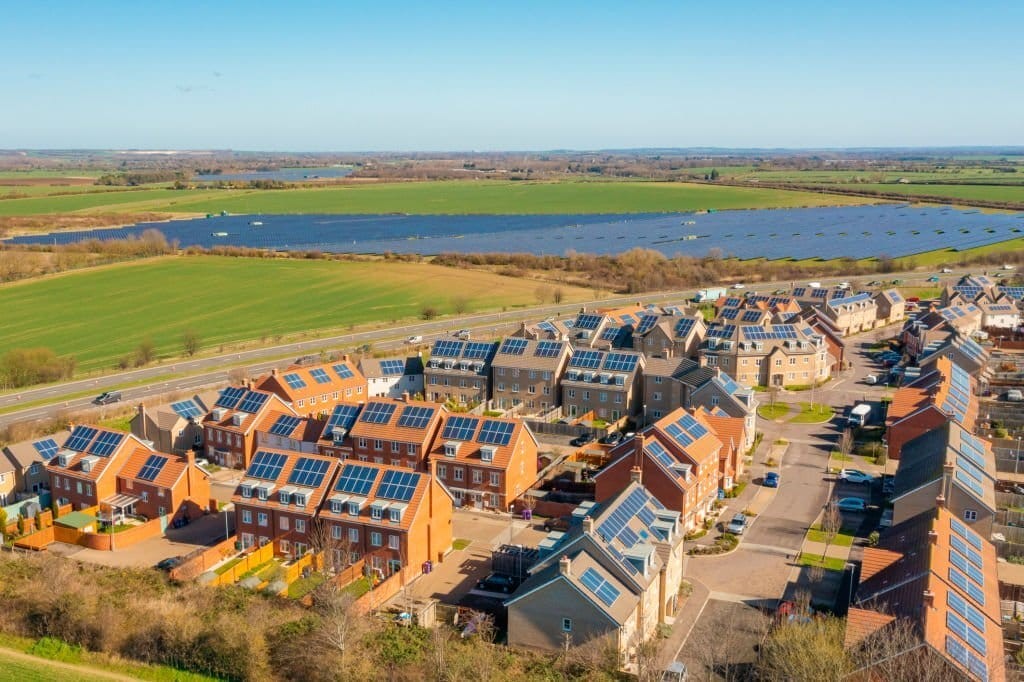 Aerial view of an eco-friendly residential development with solar panels on rooftops, bordering open green fields and a large body of water, reflecting modern sustainable living in a rural environment.