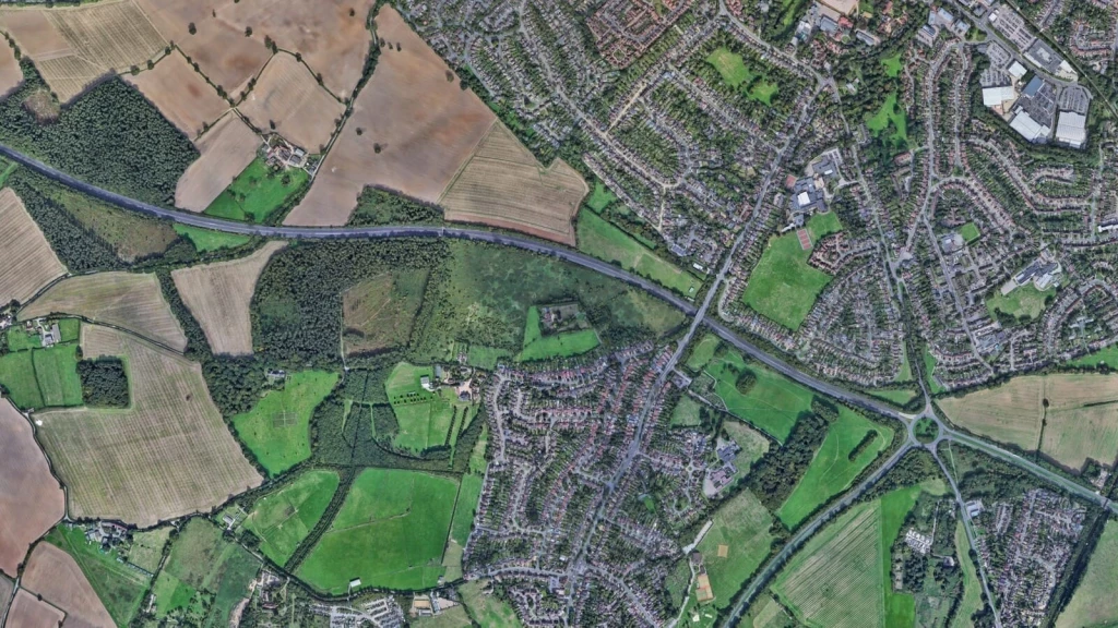 Aerial view of a countryside area showing a clear boundary between developed land with houses and expansive green fields, representing the key purposes of the Green Belt in urban planning. The image illustrates the importance of the Green Belt in preventing urban sprawl, maintaining the separation between towns, and safeguarding the countryside from encroachment. This visual is pertinent to discussions on the designated roles and purposes of Green Belt land in preserving rural landscapes and controlling urban expansion.