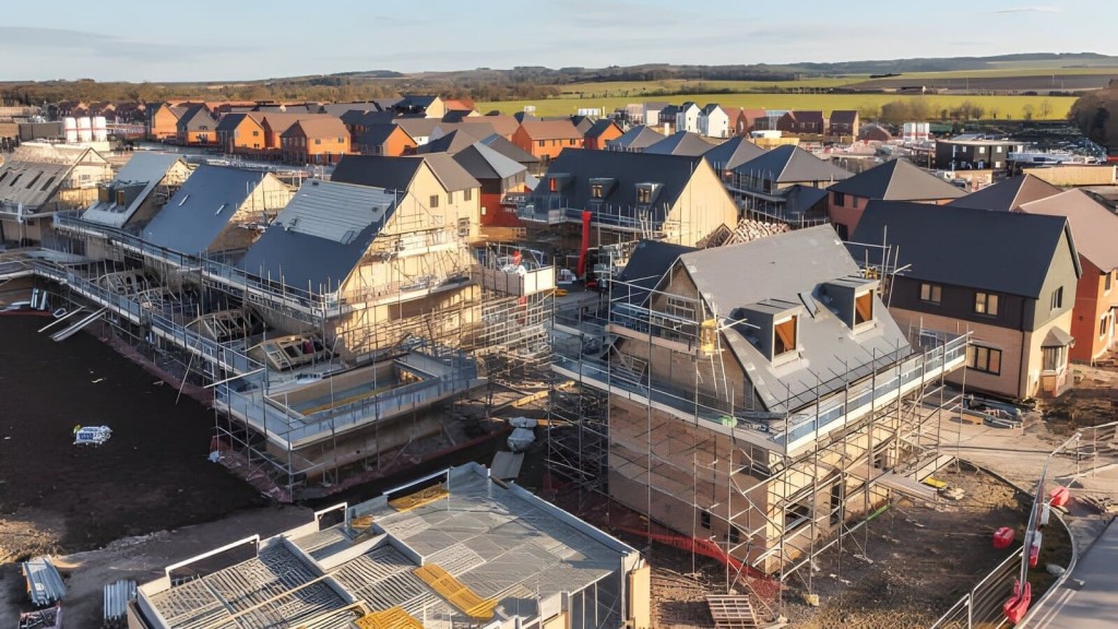 Aerial view of new housing development under construction with scaffolding around partially completed houses, located near open green fields, demonstrating expansion into Green Belt areas to address housing demands.