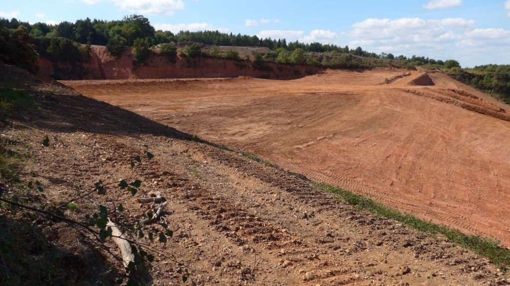 Large-scale excavation site showing exposed reddish soil and deep cuts into the landscape, surrounded by forested areas, highlighting the environmental impact of construction on Green Belt land.