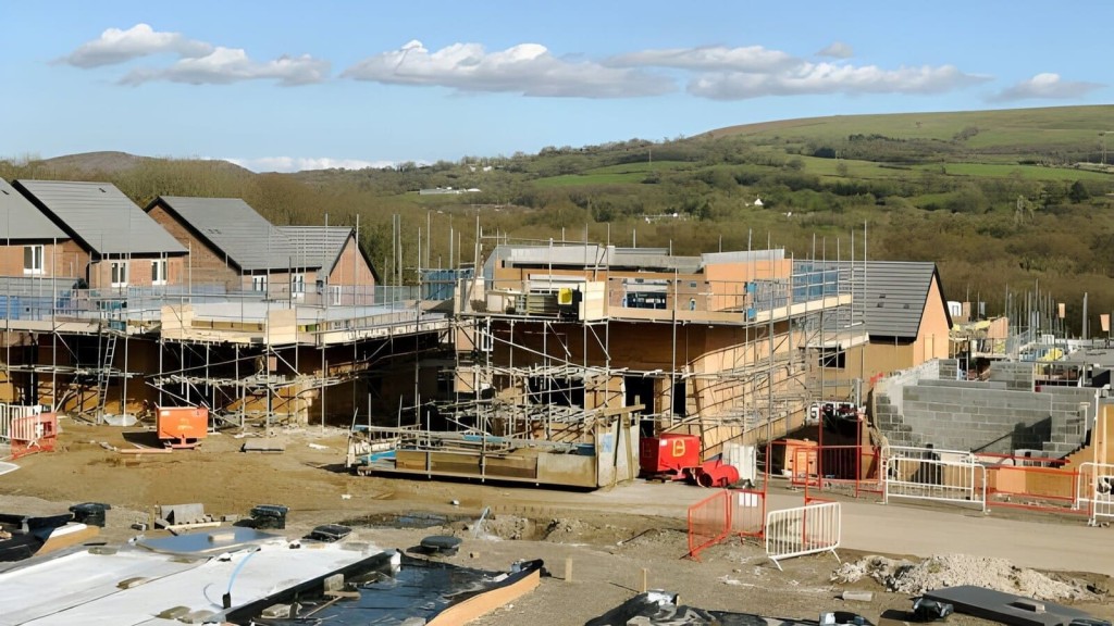 Construction site of new residential homes with scaffolding and building materials visible, set against a backdrop of rolling hills and countryside, illustrating development in rural or semi-rural settings.