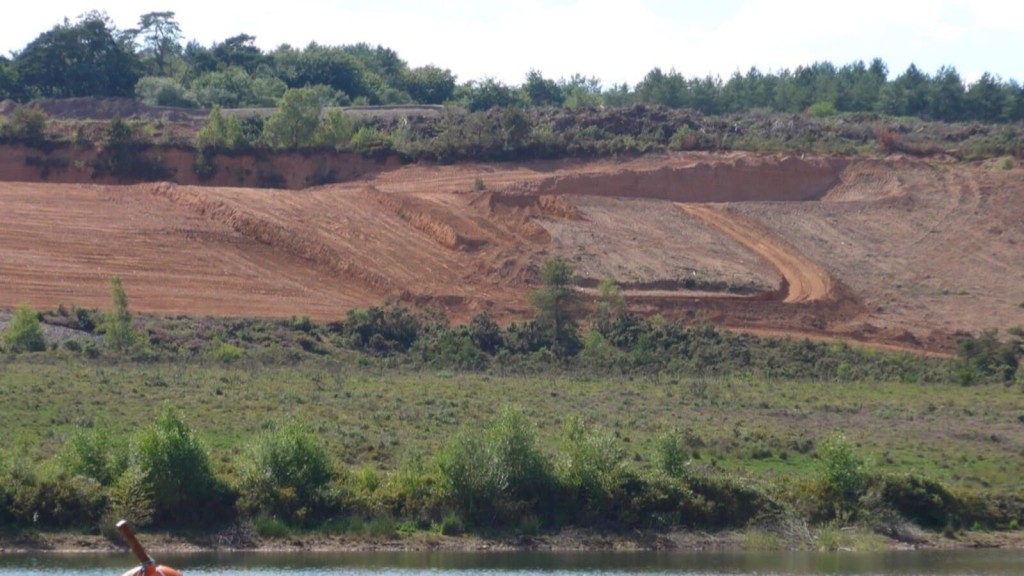 Erosion and land modification in Green Belt area showing exposed soil layers with a backdrop of lush greenery and a calm river in the foreground, illustrating environmental impact.