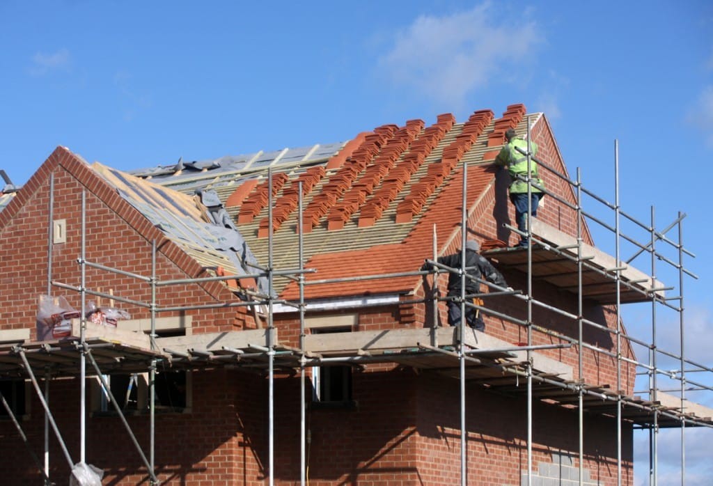 Construction workers on scaffolding installing roof tiles on a new brick house under a clear blue sky, showcasing residential construction in progress.