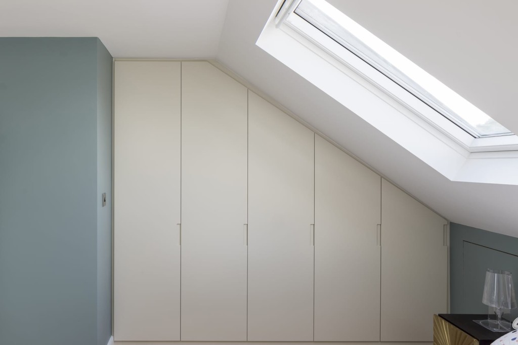 Modern minimalist interior design of a loft extension featuring sleek white built-in wardrobes under a sloped ceiling with a skylight, offering natural lighting in a stylish home.