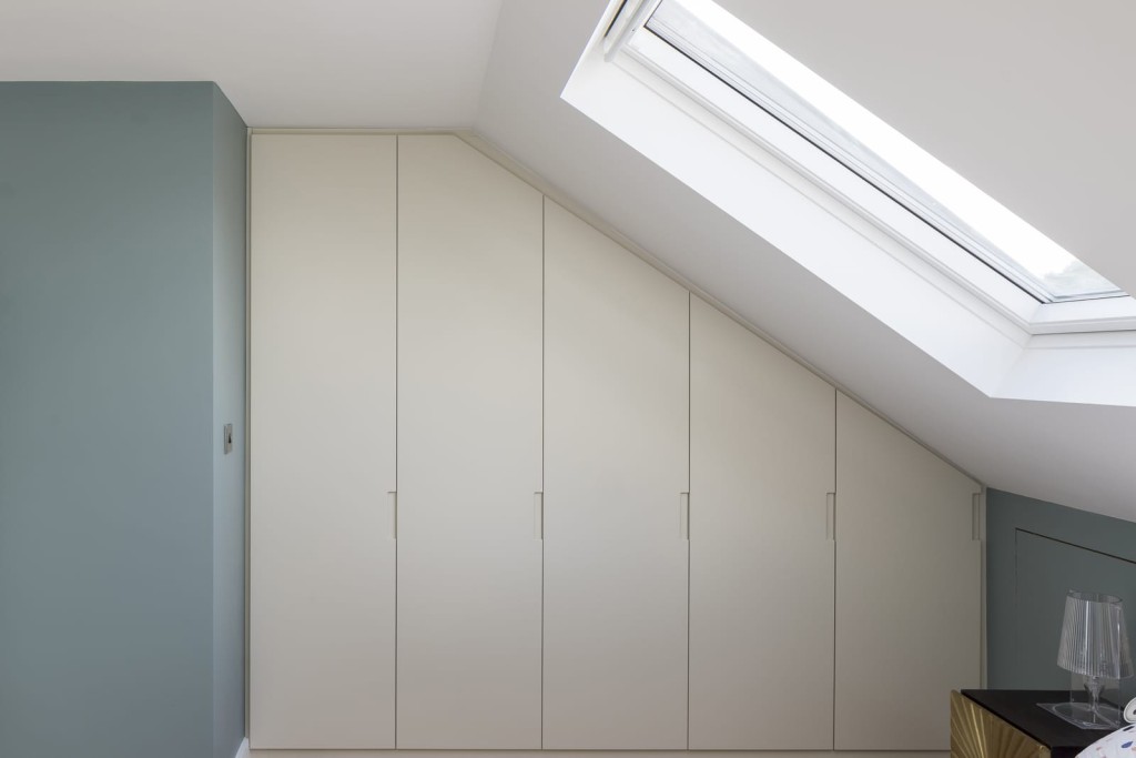 Modern and bright dormer loft extension with a large pitched roof window allowing a light of sunlight into a white and dusty blue bedroom with customised floor to ceiling wardrobe storage