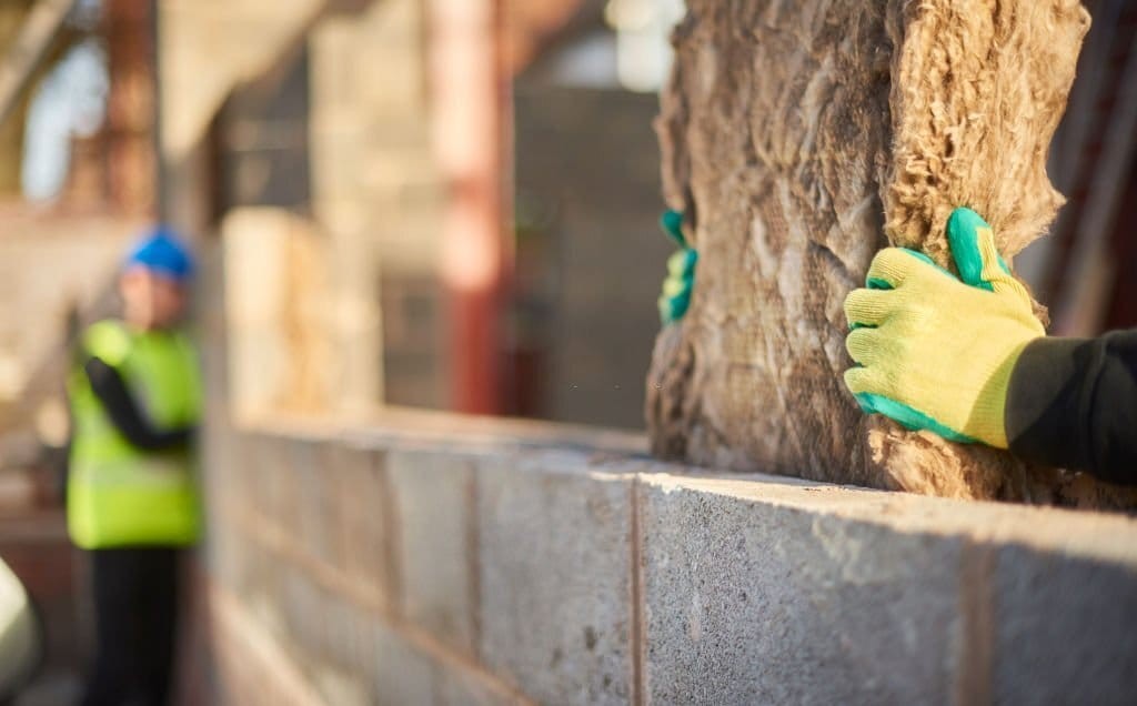Focused image of a construction worker's hands with protective gloves placing insulation materials on a brick wall, with another worker overseeing the work in the blurred background.