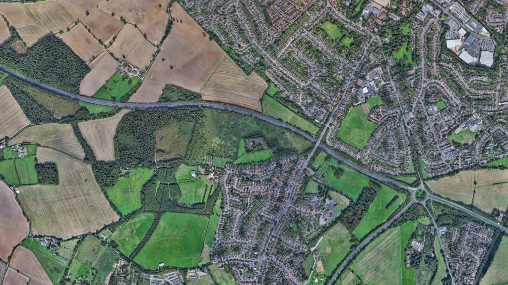 Aerial view of a suburban area bordered by agricultural fields and green spaces, illustrating the application of the 'five golden rules' for Green Belt reform. The image shows a blend of developed residential neighbourhoods and open land, highlighting the potential for sustainable housing development in underutilised 'grey belt' areas, as part of Labour's plan for efficient use of the Green Belt.