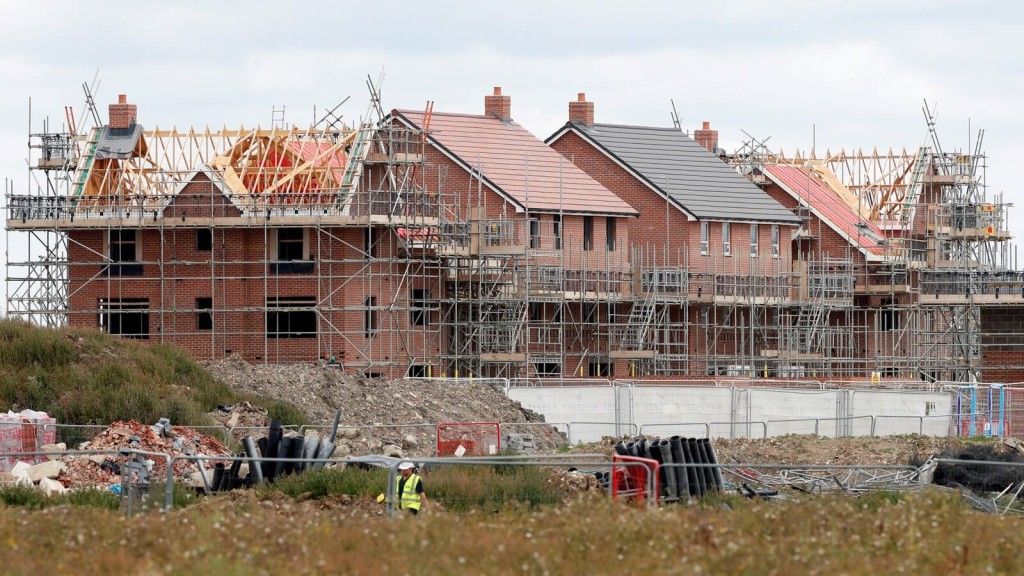 Construction site of new residential houses under development, featuring multiple homes in various stages of completion with exposed brickwork and timber roofing frames. The image includes construction workers and scaffolding, showcasing ongoing building work in a suburban development zone, highlighting urban expansion and residential construction in response to housing demand in the UK.