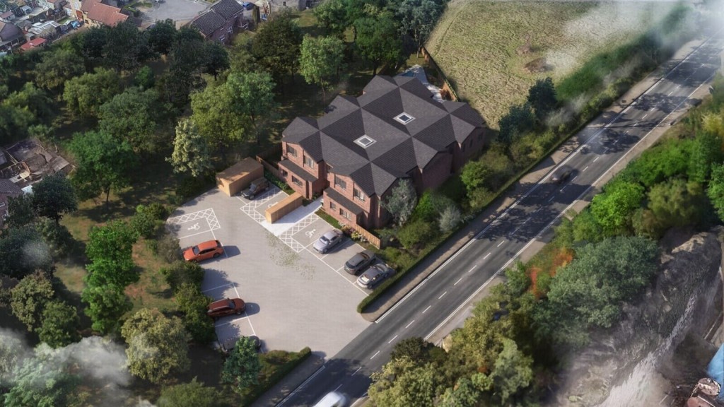 Overhead view of a modern brick residential complex with a hexagonal roof design, adjacent parking lot, and bordering green space, positioned next to a busy roadway showcasing rural planning within the green belt.