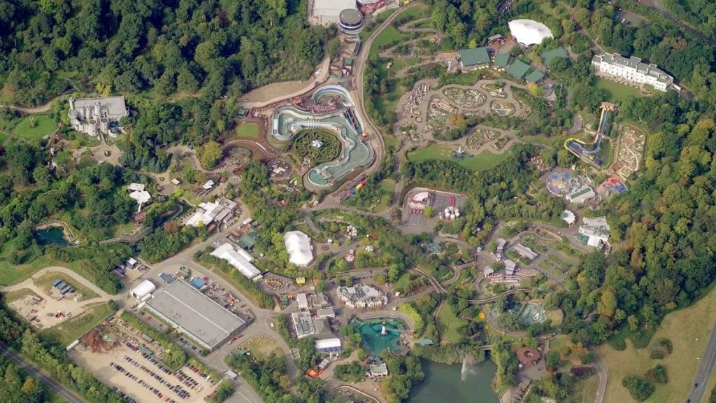 Bird's-eye view of a sprawling Legoland theme park nestled in a lush forest, featuring a variety of attractions including a water slide, roller coasters, and themed zones with meticulous landscaping, highlighting recreational development within the green belt.