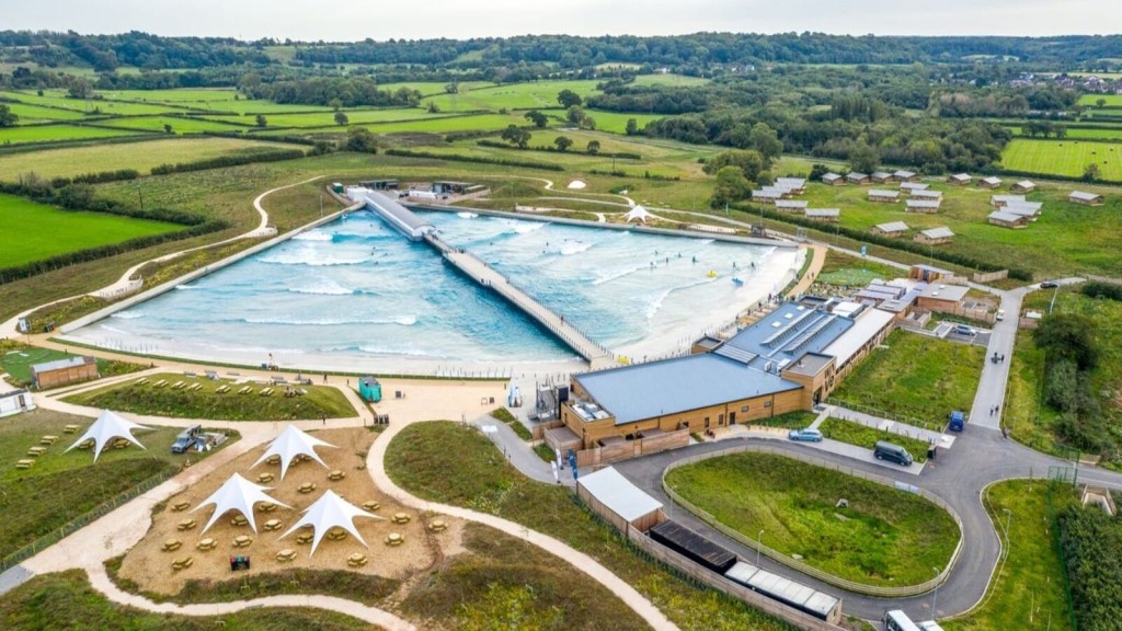 Aerial view of a modern artificial wave pool and surf center in Bristol located in a lush green landscape, showcasing the expansive, rectangular wave-generating pool with surfers, surrounded by walking paths, white canopy tents, and a main building with adjacent parking.