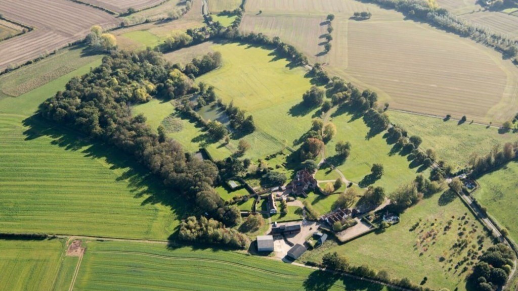 Aerial view of a green landscape showing the intricate layout of an English village surrounded by patterned agricultural fields, with a dense cluster of trees and a pond, emphasising sustainable land use in green belt areas.