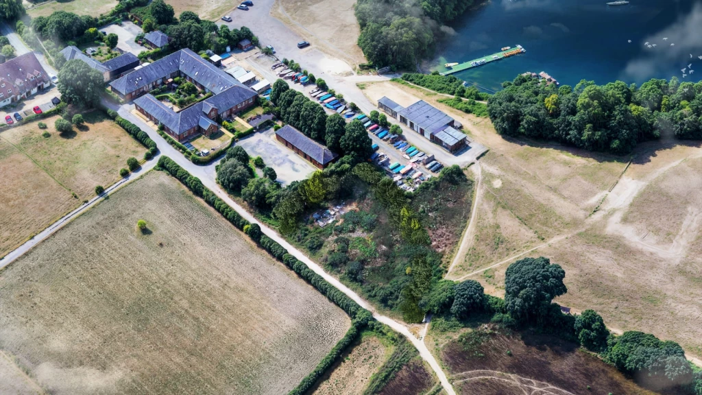 Aerial view of a Grey Belt site with existing buildings, parking areas and access tracks beside a lake, surrounded by open fields, hedgerows and trees.