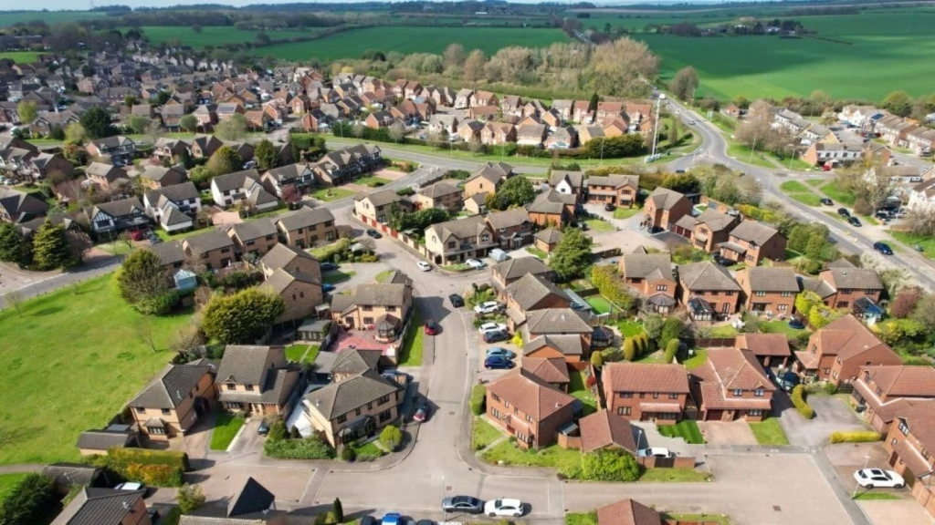 Aerial view of detached houses along residential streets, with open green space and agricultural fields beyond the settlement edge.