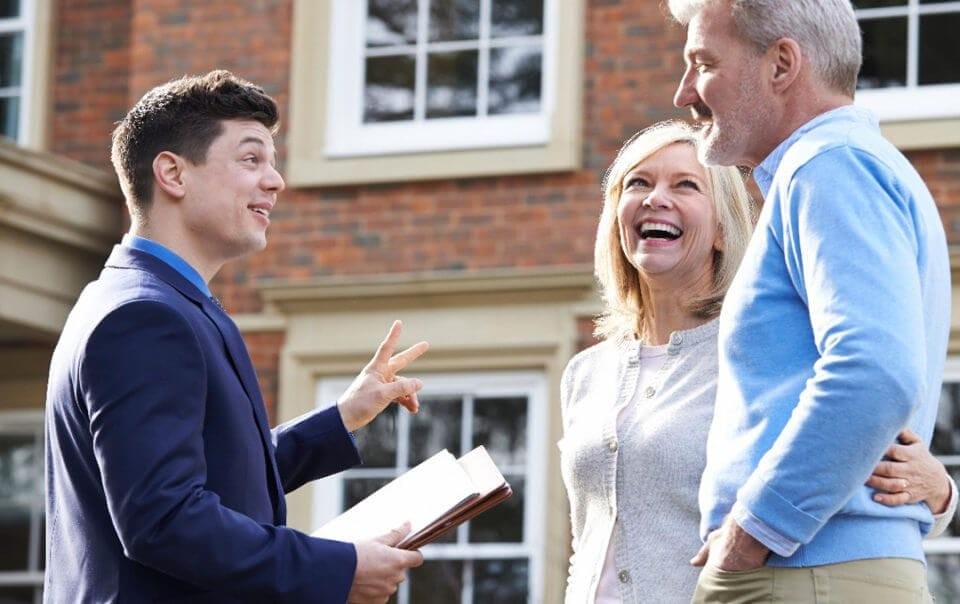 Estate agent in a two piece navy suit holding two fingers up with his left hand and paperwork on his right hand speaking to a happy and smiling couple that are holding each other around the waist outside of a large and luxurious red brick property