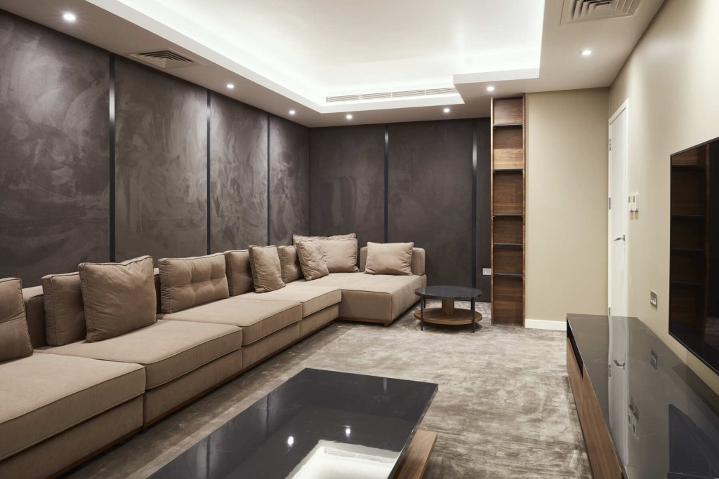 Modern living room tray ceiling with integrated spotlights, a soft suede grey wall, a very large and long beige couch with matching brown bookshelf and tv table as well as matching luxurious suede carpet just a few shades lighter