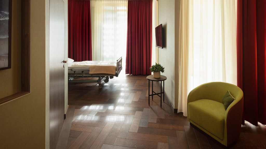 A modern hospital patient room featuring a comfortable bed, natural light streaming through large windows with elegant red curtains, and a cosy green armchair. The warm wooden flooring and thoughtful interior design create a serene, healing environment. This design prioritises patient comfort, mental wellbeing, and infection control, reflecting the key principles of healthcare architecture.