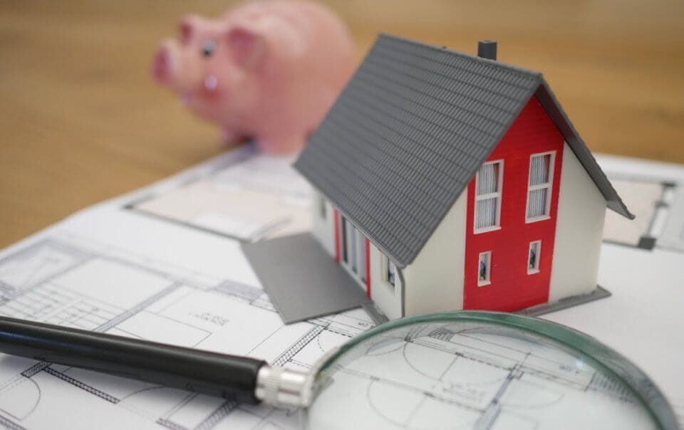 Printed technical drawing layout laying on a wooden table with a magnifying glass, a small plastic white, red and grey double storey house and a guinea piggy bank sprawled over it
