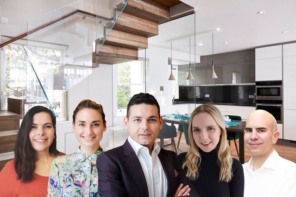 Team of architects and designers standing in a modern, stylish kitchen and living space. The background features a sleek glass staircase, contemporary kitchen cabinetry, pendant lighting, and a dining area set for a meal. The team members, consisting of two men and three women, are smiling and dressed in professional attire, ready to assist with house extension projects. This image represents the expert team at Urbanist Architecture, showcasing their commitment to high-quality, innovative home design solutions.