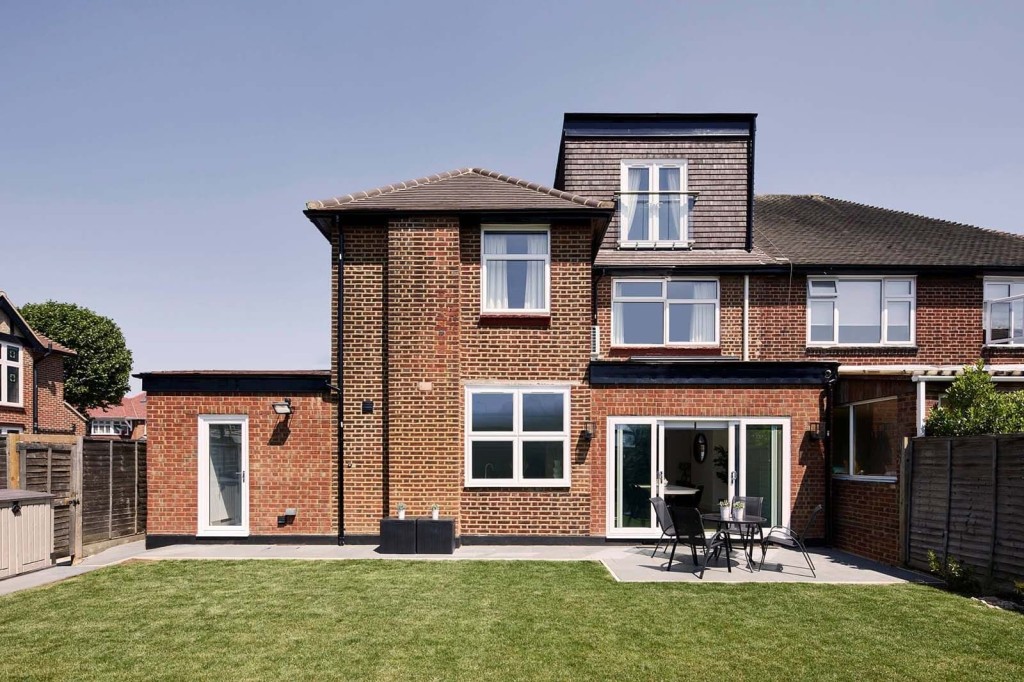 Rear view of a brick house with a modern single-storey side and loft extension and large windows and glass sliding doors, leading to a neatly manicured lawn and patio area with outdoor seating. This image illustrates a successful home extension in London, showcasing the seamless integration of new and existing structures, and highlighting the added living space and enhanced outdoor connectivity.