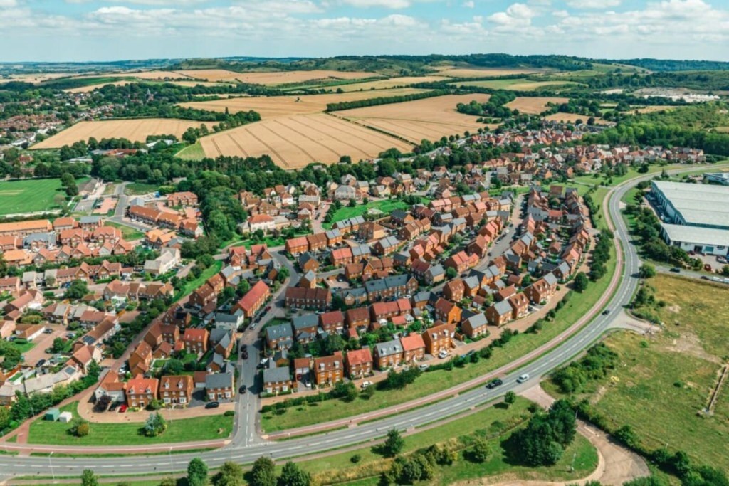 Aerial view of a vibrant residential area in the UK showcasing a variety of houses with red roofs, verdant gardens, and surrounding open fields, illustrating suburban development and community planning.