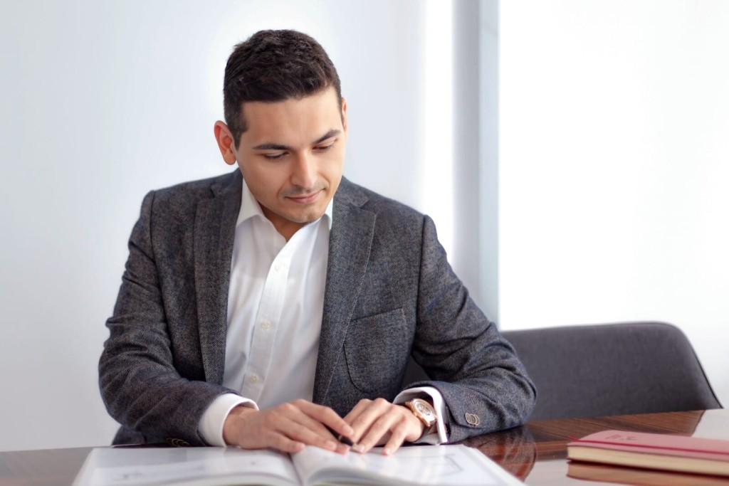 Focused young professional man, Ufuk Bahar, in a tailored grey suit and white shirt, reviewing documents at a modern office setting, exemplifying dedication and attention to detail in the workplace.