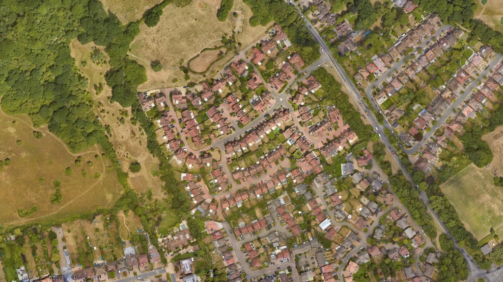 Aerial view of a UK residential neighbourhood, showing houses with private gardens arranged along streets and cul-de-sacs beside woodland and open fields. The image highlights the relationship between existing homes and surrounding green space, providing context for the challenge of increasing housebuilding while responding to the UK’s severe housing shortage.